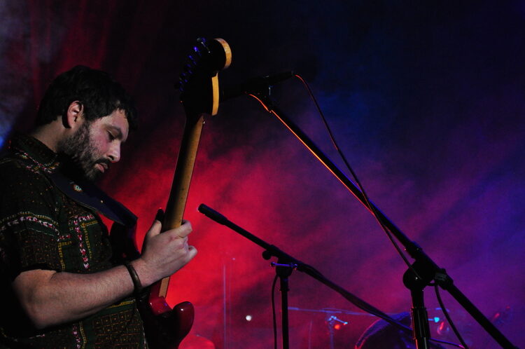 An image of Adam Carpinelli playing live on his guitar during a live show with a red/purple background.