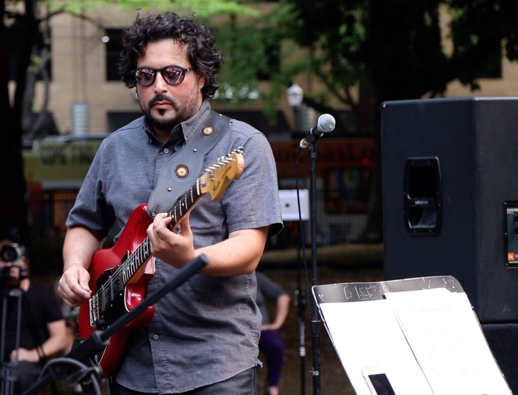 Producer and musician, Adam Carpinelli, playing on his guitar during a live show in the park.